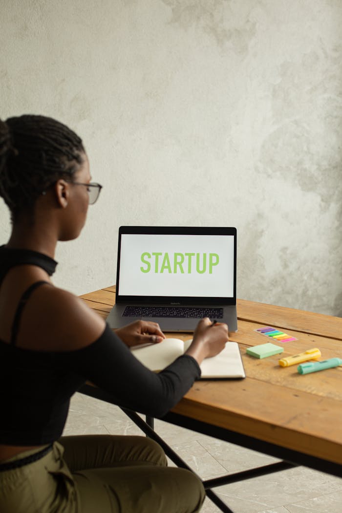 Side view of anonymous African American female entrepreneur sitting at table with netbook with startup inscription on screen and writing in notebook using pen and markers
