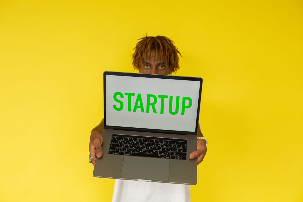 about-01 A young black man holds a laptop displaying 'Startup' against a vibrant yellow background.